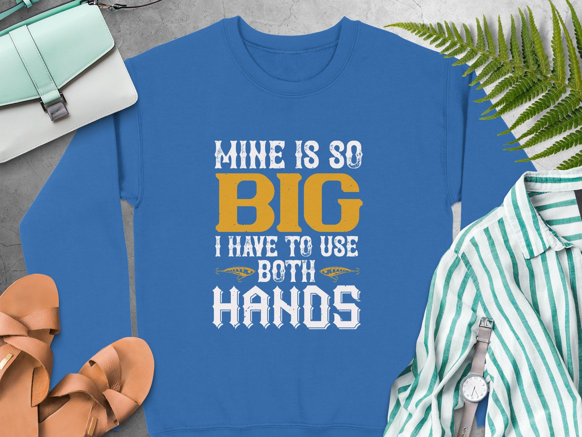A vibrant blue sweatshirt by Garment Graphics boldly says, MINE IS SO BIG I HAVE TO USE BOTH HANDS. It sits amidst a striped shirt, brown sandals, a wristwatch, and a green bag on ferns against a gray backdrop.