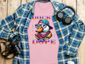 A pink Garment Graphics T-Shirt, featuring a cartoon duck with glasses and a cap, lies on wood. Its unique style pops among a plaid shirt, headphones, camera, and coffee cup. The Duck Dope text adds to its eye-catching design.