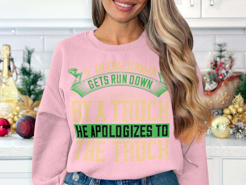 A woman with long blonde hair wears a bold, humorous pink sweatshirt by Garment Graphics featuring green and yellow text: If an Englishman gets run down by a truck, he apologizes to the truck. It fits perfectly as she stands in a kitchen with holiday decorations.