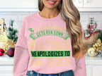 A woman with long blonde hair wears a bold, humorous pink sweatshirt by Garment Graphics featuring green and yellow text: If an Englishman gets run down by a truck, he apologizes to the truck. It fits perfectly as she stands in a kitchen with holiday decorations.