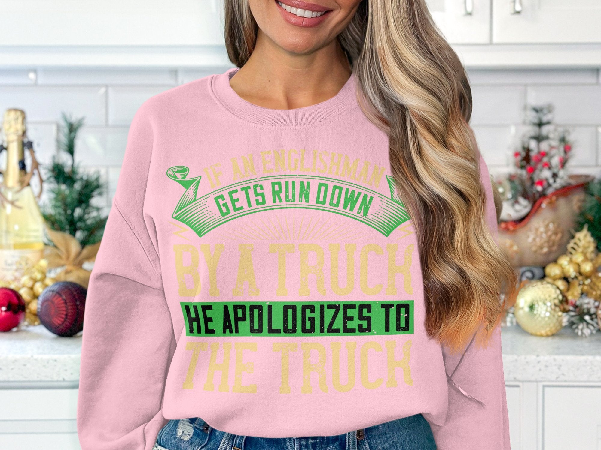 A woman with long blonde hair wears a bold, humorous pink sweatshirt by Garment Graphics featuring green and yellow text: If an Englishman gets run down by a truck, he apologizes to the truck. It fits perfectly as she stands in a kitchen with holiday decorations.
