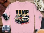 A pink sweatshirt from Garment Graphics with a rally car graphic and YUMP on it is laid out on wood. Nearby items include a camera, leather gloves, binoculars, a carabiner, and a strap. This design captures the thrill of adventure and style in one piece.