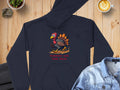 A black Garment Graphics hoodie featuring the Turkey on the Run graphic is placed on a wooden surface, accompanied by a coffee cup, a small plant, a denim jacket, and part of another dark garment for a cozy vibe.