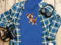 This Garment Graphics blue hoodie features a cartoon puppy print and is presented on a wooden surface. Nearby are a checkered blue and green shirt, black headphones, a camera for capturing moments, and a disposable cup with white lid—perfect additions to any dog lover’s wardrobe!.