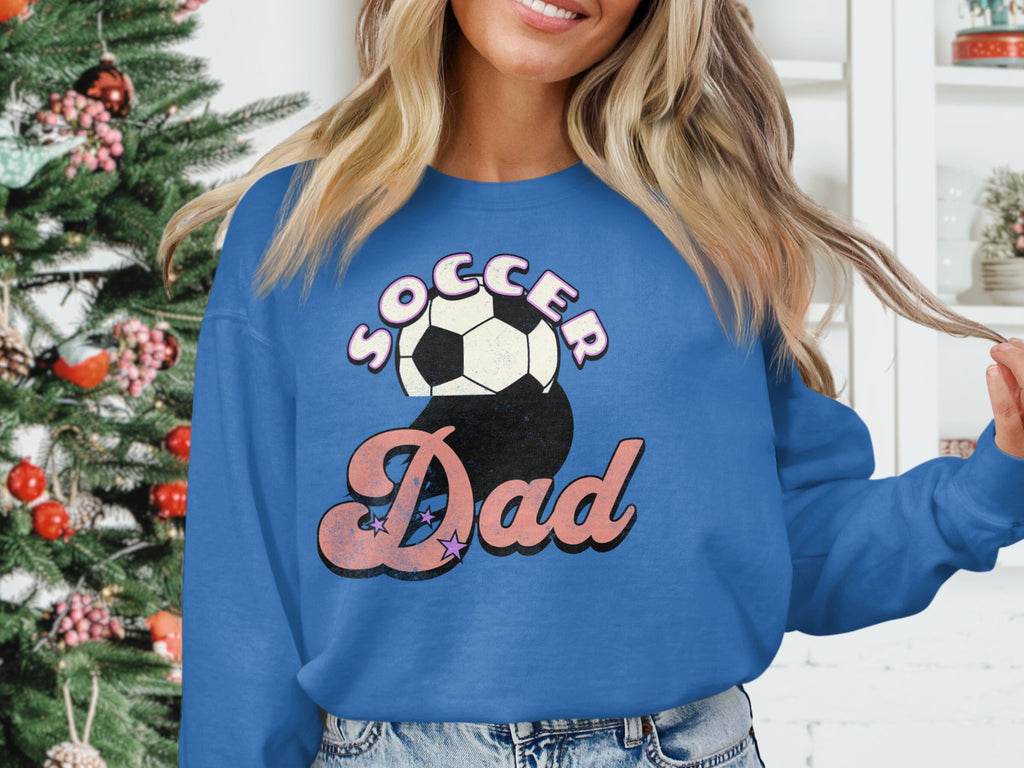 A person wearing a Garment Graphics blue sweatshirt with a Soccer Dad retro design and soccer ball stands indoors. A beautifully decorated Christmas tree with ornaments is in the background, alongside a white shelf displaying various objects.