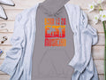 A gray Unisex Fashion Hoodie by Garment Graphics features an orange and red Born to Be Musician chest graphic with silhouettes of music lovers. Styled on wood, its paired with a gray cardigan, flowers, and accessories for the perfect ensemble.