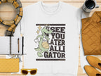 The Garment Graphics white alligator graphic sweatshirt showcases a cartoon alligator with a basketball and See You Later Alligator text, styled on a wooden surface with a yellow scarf, sunglasses, a notebook, pen, quilted jacket, and coffee cup.