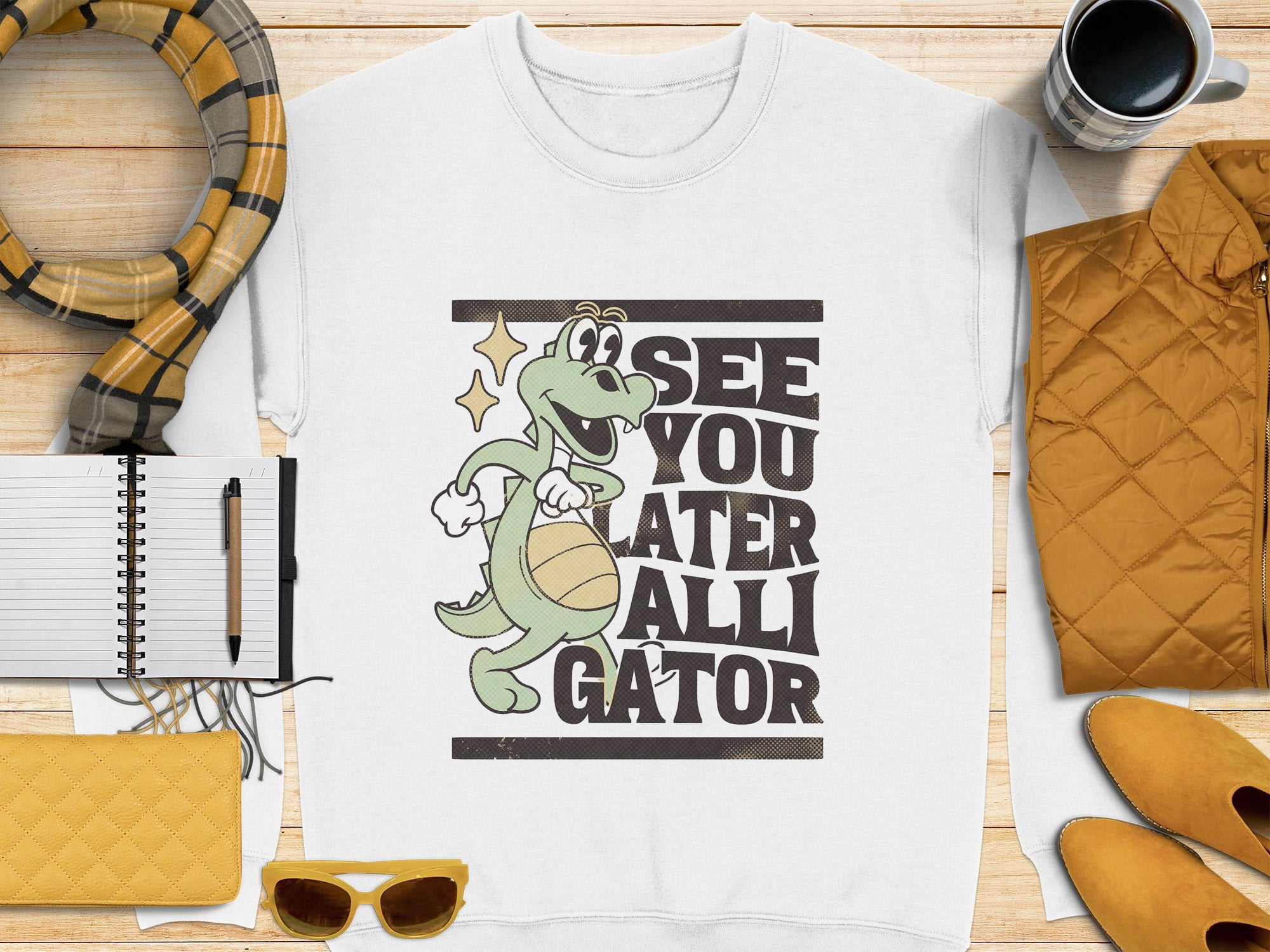 The Garment Graphics white alligator graphic sweatshirt showcases a cartoon alligator with a basketball and See You Later Alligator text, styled on a wooden surface with a yellow scarf, sunglasses, a notebook, pen, quilted jacket, and coffee cup.
