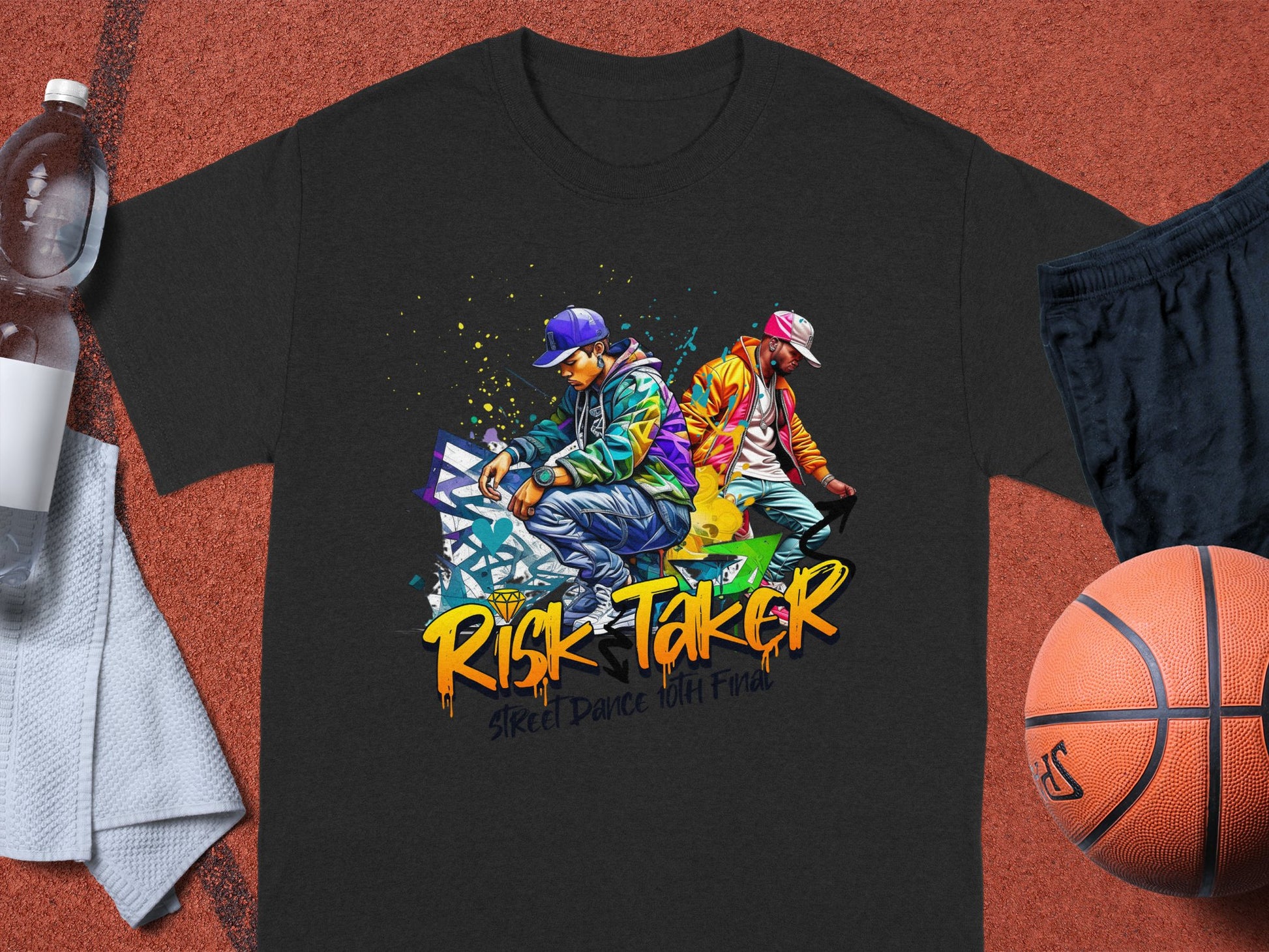 Garment Graphics black T-shirt showcases two street dancers, one in blue and the other in red-yellow. The text Risk Taker: Street Dance with Fire celebrates street dance culture. Featured with a water bottle, gray towel, and basketball.