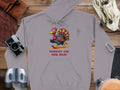 Garment Graphics gray hoodie showcases a cartoon turkey running with Turkey On The Run in bold red. Its displayed on wood, surrounded by essentials like a camera, binoculars, gloves, carabiner, and a leather pouch.