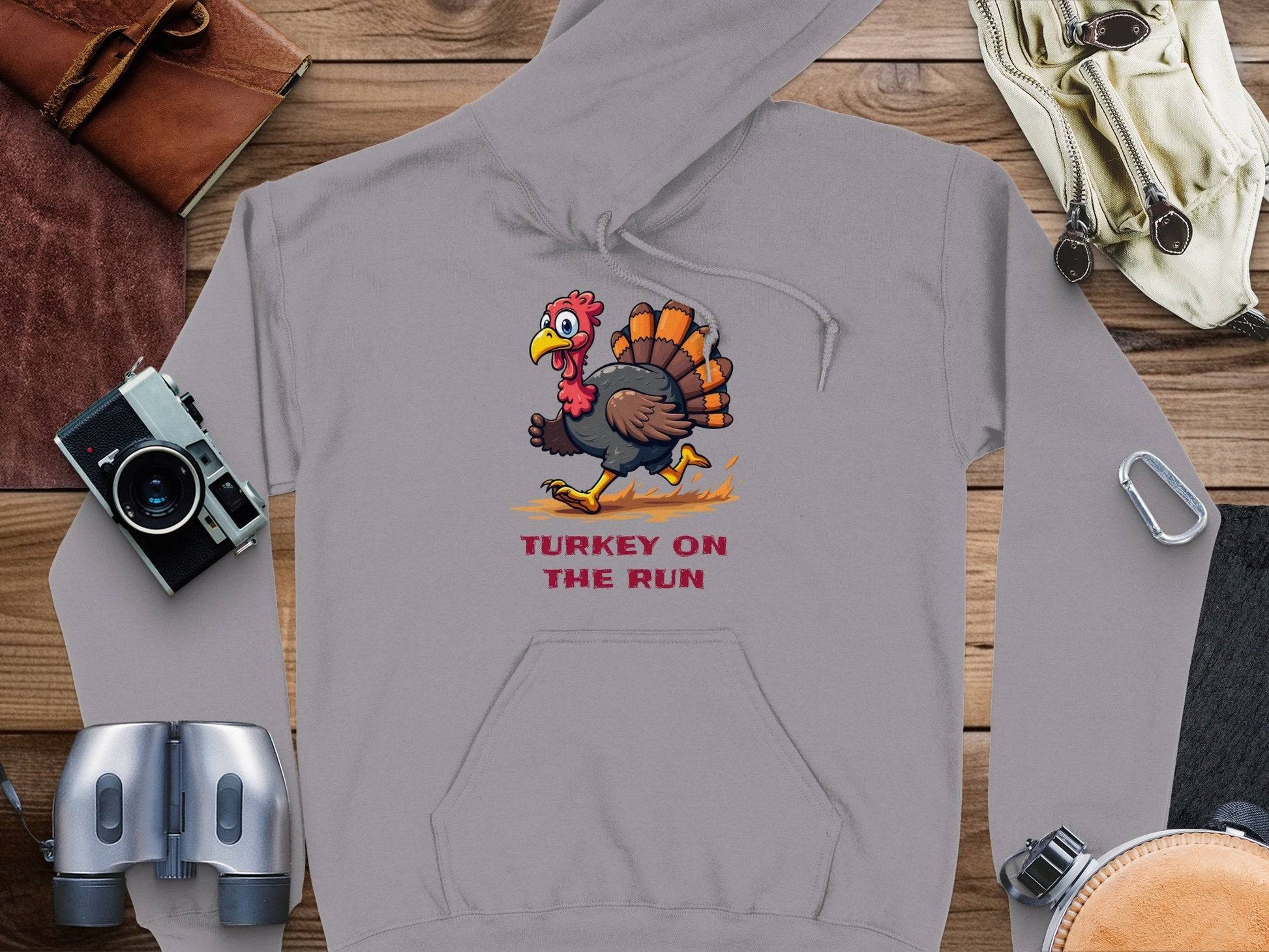 Garment Graphics gray hoodie showcases a cartoon turkey running with Turkey On The Run in bold red. Its displayed on wood, surrounded by essentials like a camera, binoculars, gloves, carabiner, and a leather pouch.