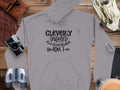 A Garment Graphics gray Cleverly Disguised as a Responsible Adult hoodie is laid flat on wood, alongside a camera, binoculars, leather pouch, and gloves—ideal for anyone embracing stylish casual wear.