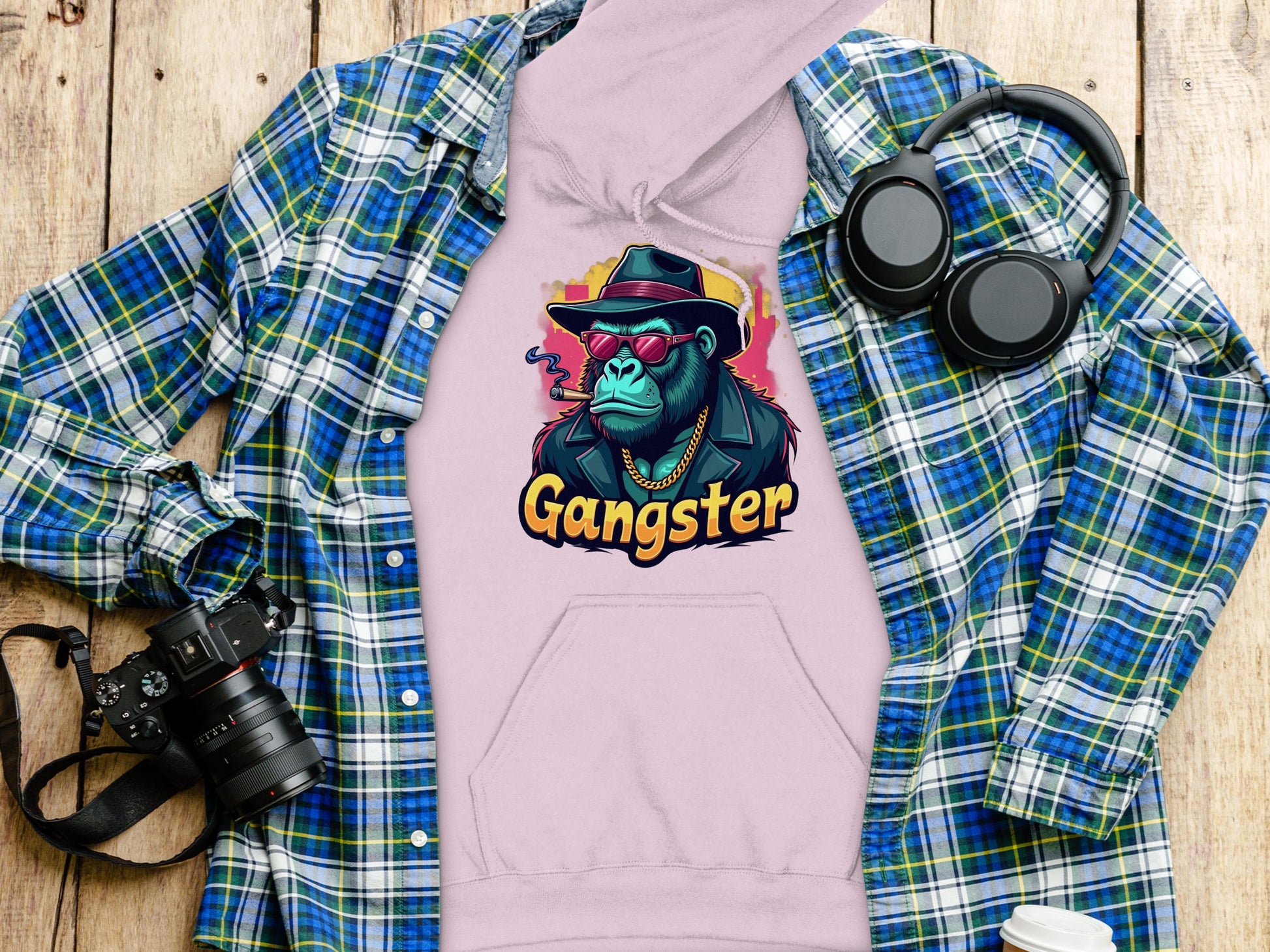 A flat lay showcases a pink hoodie by Garment Graphics, featuring a graphic of a gangster ape in sunglasses and a hat labeled Gangster. Surrounded by a plaid shirt, headphones, camera, and coffee cup on wooden surface, this piece embodies hipster urban style.