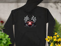 This Garment Graphics black hoodie, featuring a red racing car and checkered flags graphic, is displayed flat among green plants and yellow flowers on concrete.