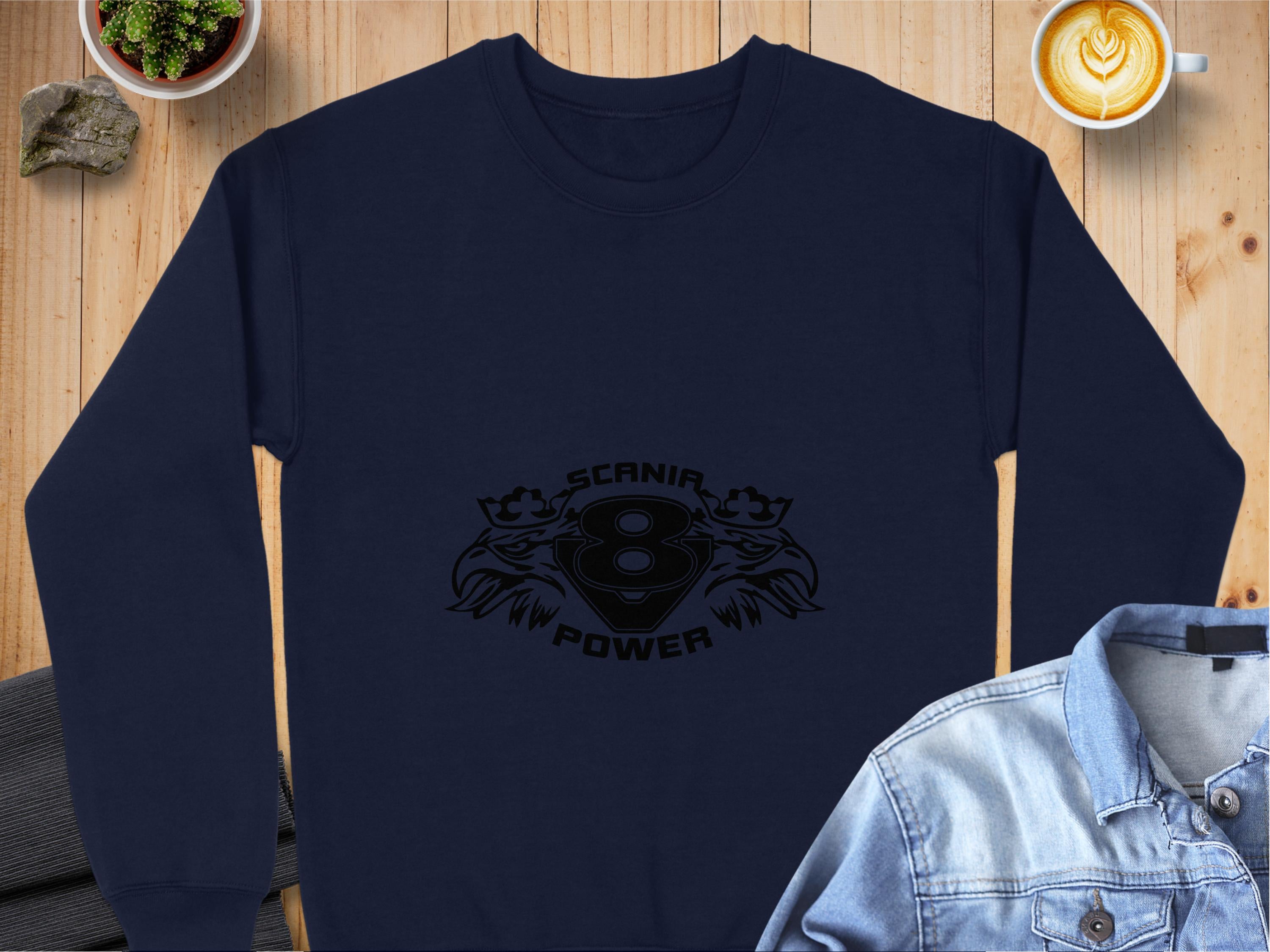 A navy blue sweatshirt from Garment Graphics showcases a sleek Scania Power V8 logo with black birds on a wooden background. Nearby are the collar of a denim jacket and a latte in a white cup.