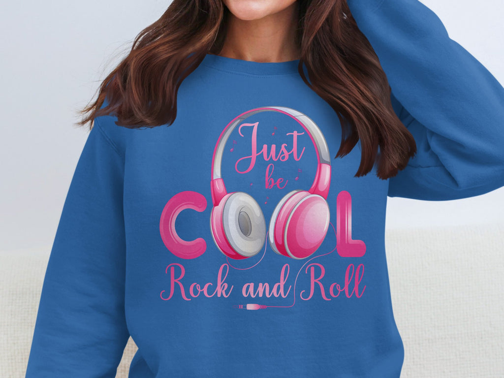 A fashion lover dons a Garment Graphics blue sweatshirt featuring pink Rock and Roll headphones framing Cool. Bright pink text says Just be Cool Rock and Roll against white, making it essential for music fans.