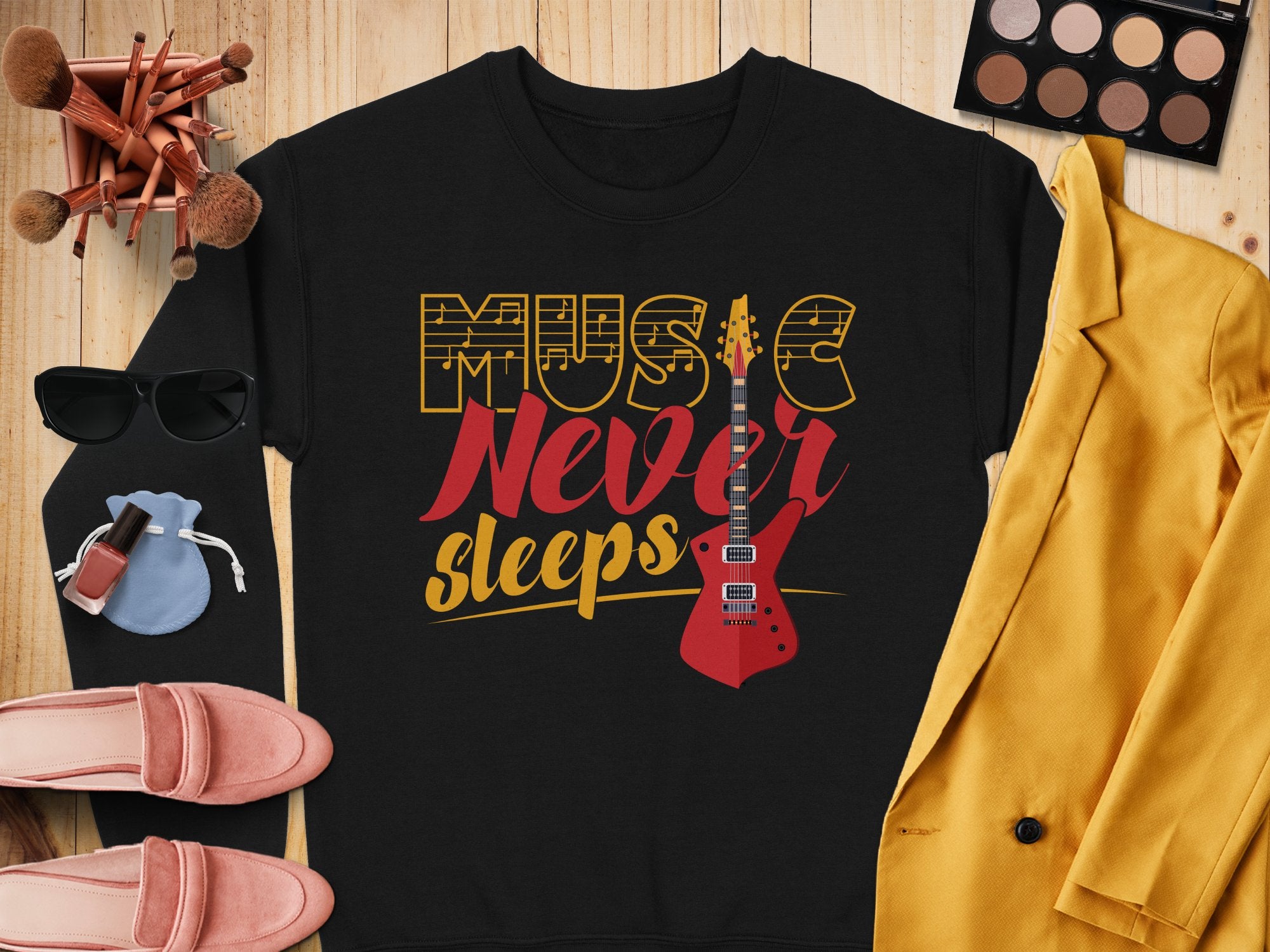 A stylish Garment Graphics sweatshirt with the Music Never Sleeps Guitar design is displayed on a wooden surface. Surrounding it are makeup brushes, an eyeshadow palette, sunglasses, a yellow jacket, a small bag, nail polish, lipsticks, and pink shoes—ideal for music lovers with fashion flair.