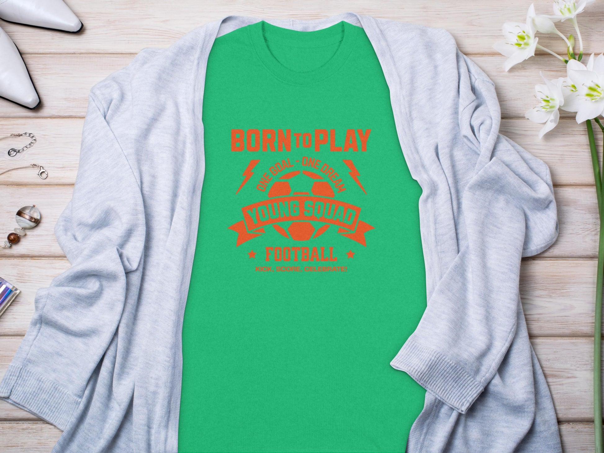 Garment Graphics green football T-shirt, with BORN TO PLAY and YOUNG SQUAD FOOTBALL in bold red, is displayed on wood. Paired with a gray cardigan and white flowers, it captures the essence of One Goal One Dream.