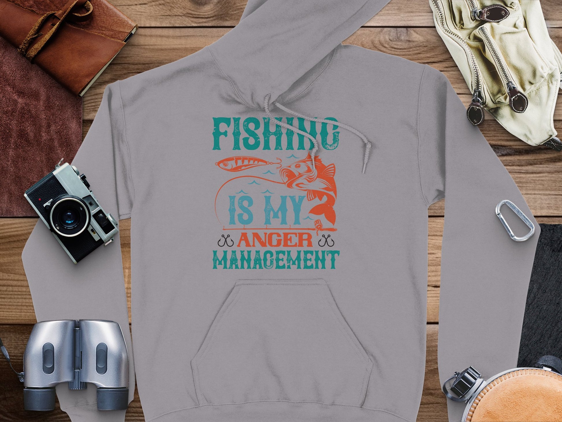 A Garment Graphics gray hoodie, perfect for an anglers wardrobe, is laid flat on a wooden surface with a camera, binoculars, headphones, watch, and leather pouch. It features a fish graphic and the text Fishing is my anger management in blue, red, and orange.