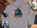 A Garment Graphics gray hoodie, exemplifying hipster urban style, displays a graphic of a gangster ape in a fedora, sunglasses, and gold chain with Gangster beneath. It is laid on wood and surrounded by travel essentials.