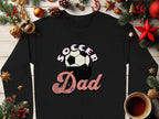 A black Garment Graphics sweatshirt with a Soccer Dad retro design and soccer ball graphic is displayed on wood. Its surrounded by festive Christmas decorations, including pine branches, ornaments, pinecones, a candy cane, and a steaming cup of coffee.