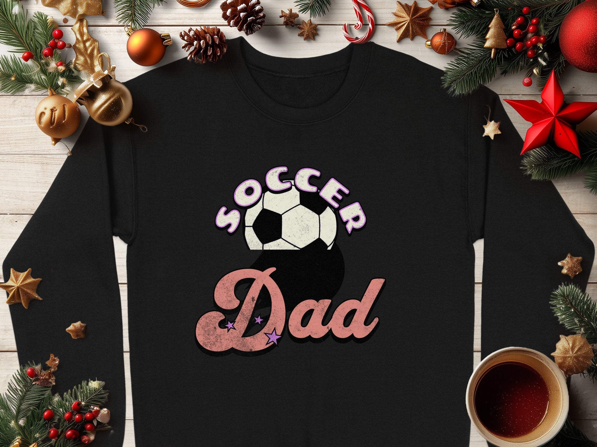 A black Garment Graphics sweatshirt with a Soccer Dad retro design and soccer ball graphic is displayed on wood. Its surrounded by festive Christmas decorations, including pine branches, ornaments, pinecones, a candy cane, and a steaming cup of coffee.