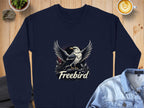 A navy Garment Graphics sweatshirt on a wooden table features an eagle with spread wings and foliage in the background. The word Freebird is boldly displayed below. Its surrounded by a denim jacket, a latte, and a small potted plant. Available in unisex sizing.