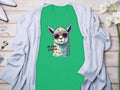 A Garment Graphics green Llama T-Shirt features an illustrated llama with red sunglasses and the caption No Prob-llama. Placed on a light wooden surface beneath a gray cardigan, it’s ideal for family sizing. Nearby, white flowers and jewelry complete the elegant setup.