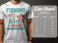 A fishing enthusiast wears a Garment Graphics t-shirt featuring Fishing Is My Anger Management with colorful text and a fish graphic. Nearby, a size chart details the dimensions for Gildan 5000 heavy cotton tees in various sizes.