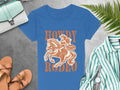 The Womens Blue T-shirt by Garment Graphics features an orange cowboy on a horse graphic with Howdy Rodeo and Not a Morning Person text. Its showcased on a gray surface with a fern, striped shirt, sandals, bag, and watch nearby—a must-have for unique Rodeo T-Shirt enthusiasts!.