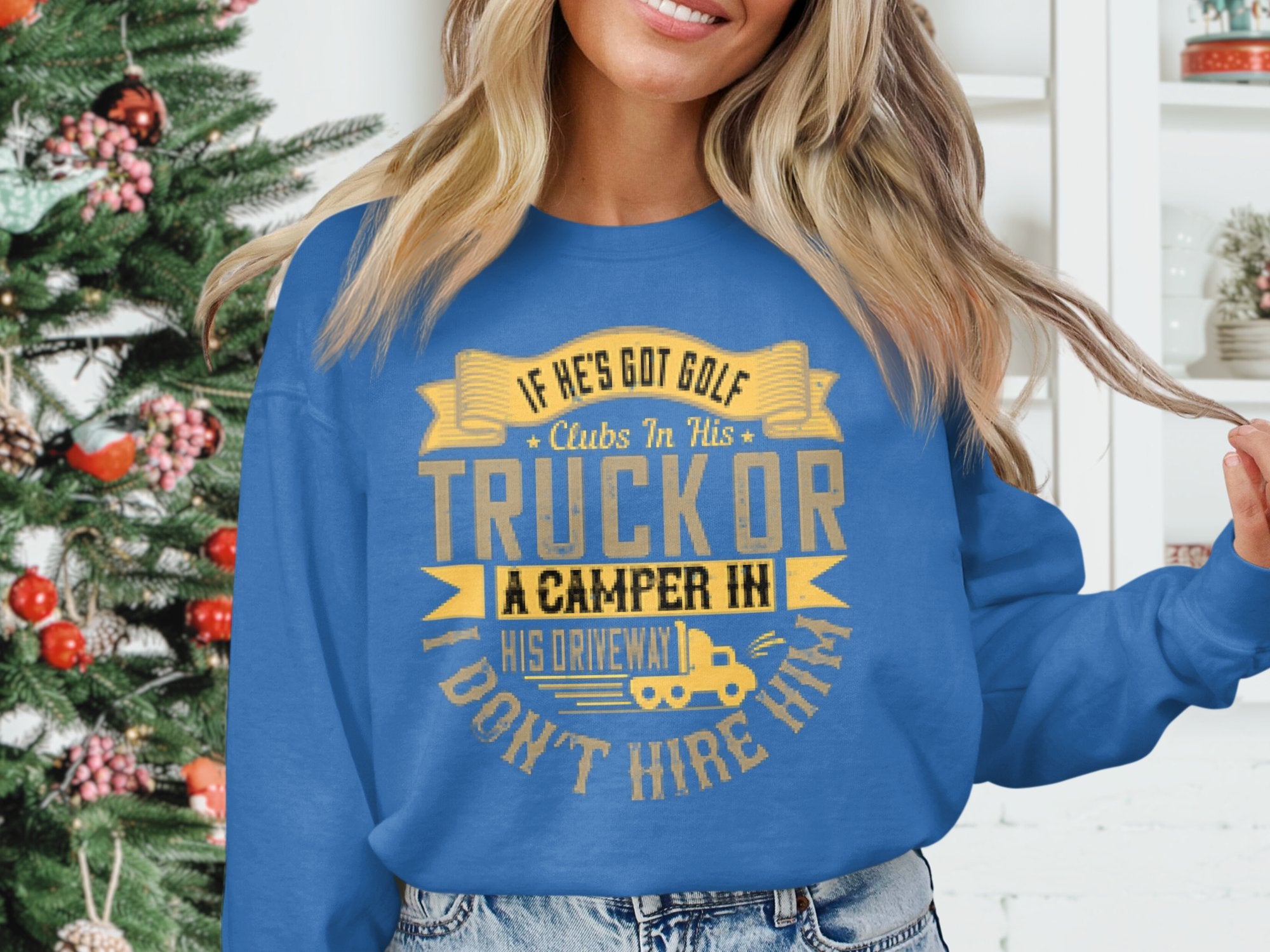 A woman wears a Garment Graphics blue sweater with the funny message: If hes got golf clubs in his truck or a camper in his driveway, I dont hire him. A decorated Christmas tree enhances the festive atmosphere, highlighting her quirky outfit.