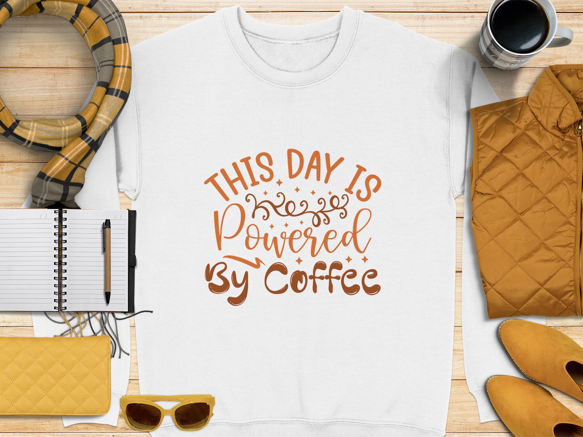 Garment Graphics presents a playful white graphic sweatshirt with This day is powered by coffee in brown lettering, surrounded by a notebook and pen, yellow scarf, steaming cup of coffee, quilted jacket, sunglasses, and a chic quilted purse design.