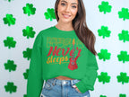 In front of a wall with green shamrock cutouts, a woman proudly wears a Music Never Sleeps Guitar sweatshirt by Garment Graphics. Her long dark hair flows as she smiles slightly, capturing the essence of music lovers everywhere.