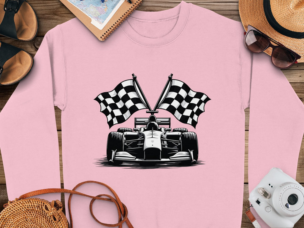 The Garment Graphics sweatshirt showcases a pink design with a black and white racing car graphic, accented by two checkered flags. Its displayed on a wooden surface alongside a hat, sunglasses, camera, sandals, notebook, and map.
