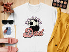 The Garment Graphics Soccer Dad sweatshirt features a retro design with vibrant purple and pink lettering alongside a soccer ball. Its displayed on wood, surrounded by a yellow jacket, pink shoes, a makeup palette, sunglasses, brushes, and a purse.