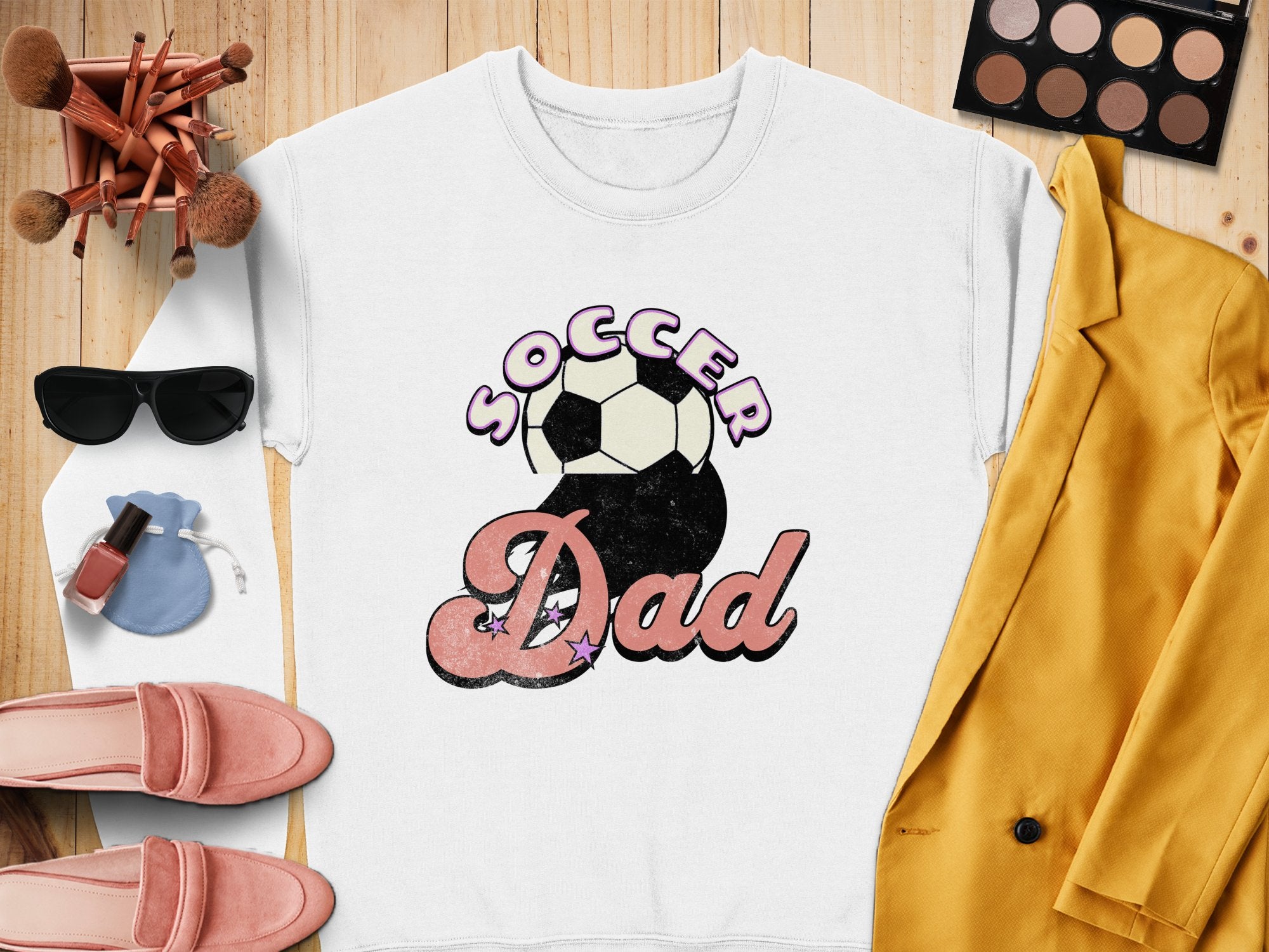 The Garment Graphics Soccer Dad sweatshirt features a retro design with vibrant purple and pink lettering alongside a soccer ball. Its displayed on wood, surrounded by a yellow jacket, pink shoes, a makeup palette, sunglasses, brushes, and a purse.