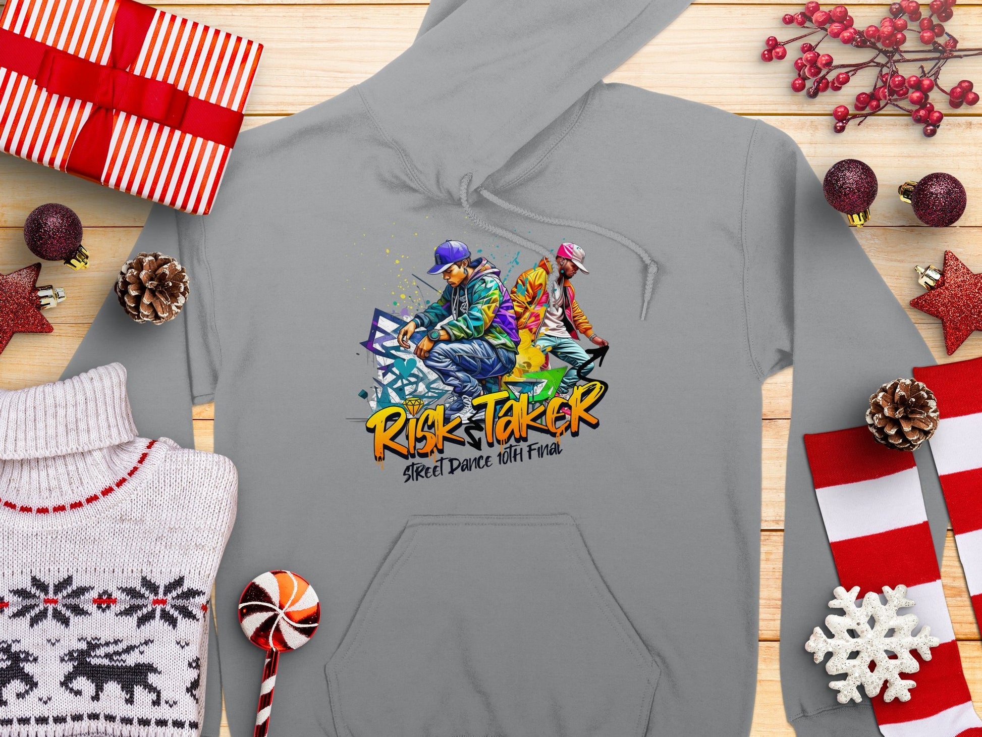 A bold gray hoodie from Garment Graphics features two dancers and Risk Taker Street Dance 10th Final. Its stylishly displayed on wood, adorned with holiday decor, gifts, pinecones, and candy canes.