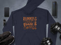 A Garment Graphics navy blue classic fit hoodie with Running on grace & coffee in orange, surrounded by a smartphone, smartwatch, black sneakers, dumbbells, and a black portable speaker on a gray surface. Made from medium-heavy fabric for comfort and style.