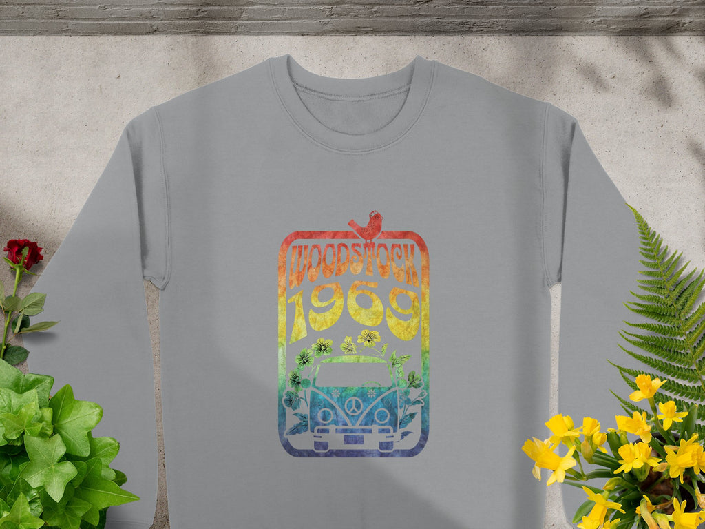 Garment Graphics gray sweatshirt features a colorful design of a Volkswagen Beetle, flowers, and a bird, all under the text Woodstock 1969, capturing the vintage spirit of the festival amidst potted plants on concrete.