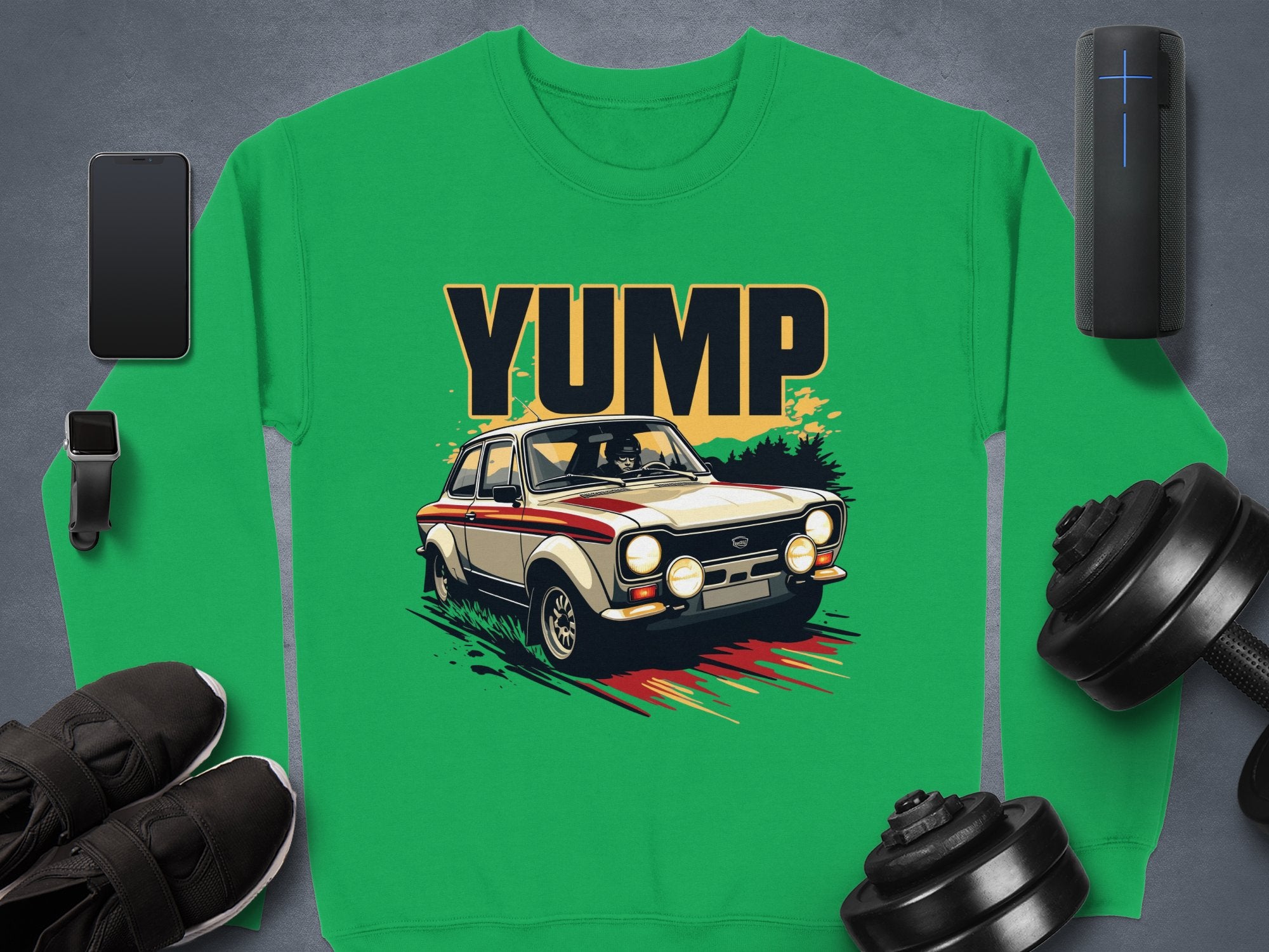 A Garment Graphics green sweatshirt features a YUMP rally car design, elegantly displayed with a smartphone, smartwatch, wireless speaker, athletic shoes, and dumbbells on a sleek gray surface.