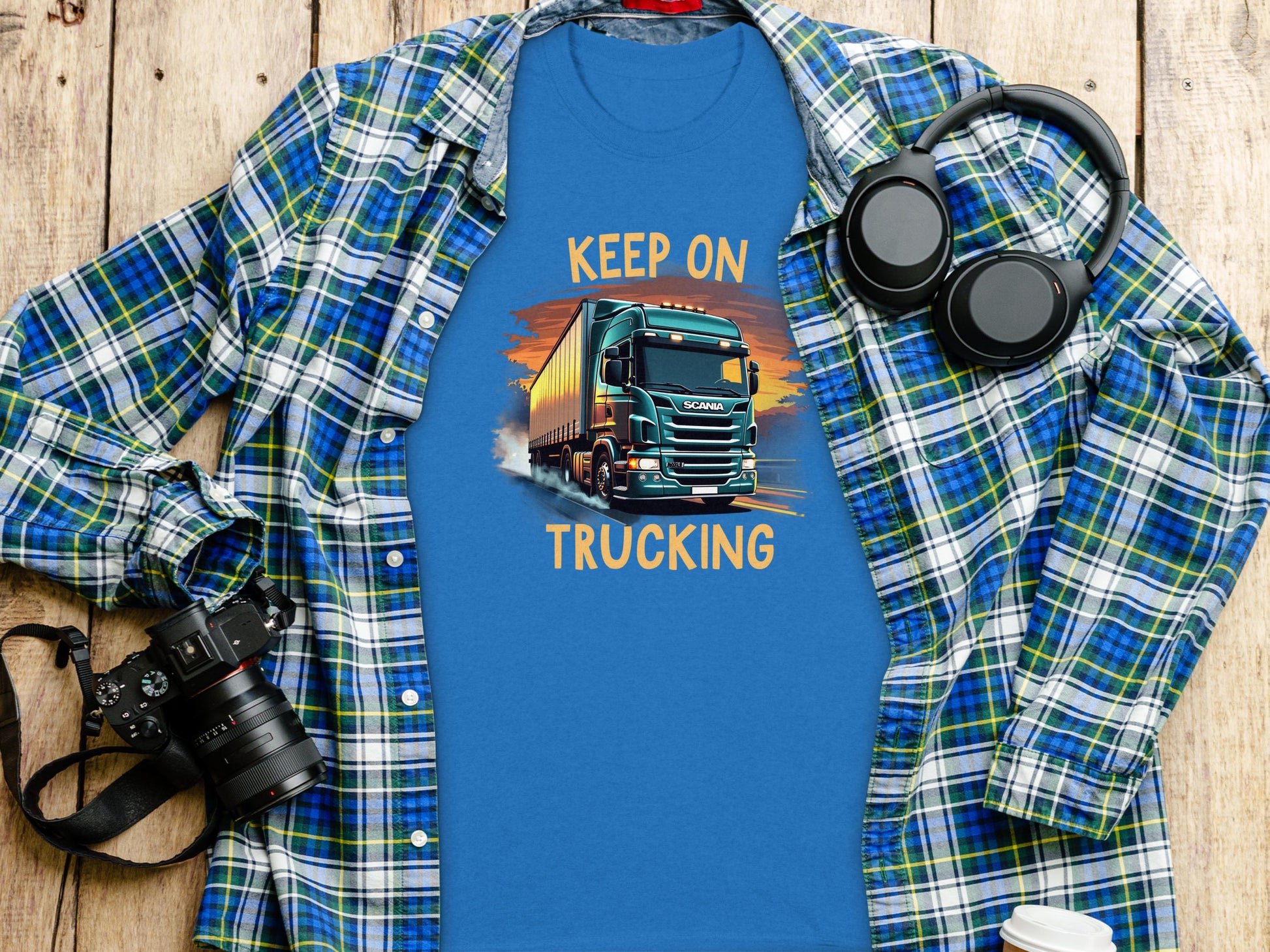 A Garment Graphics blue graphic T-shirt for truck lovers showcases Keep On Trucking with a truck image on wood. Nearby are a plaid shirt, headphones, camera, and disposable coffee cup.