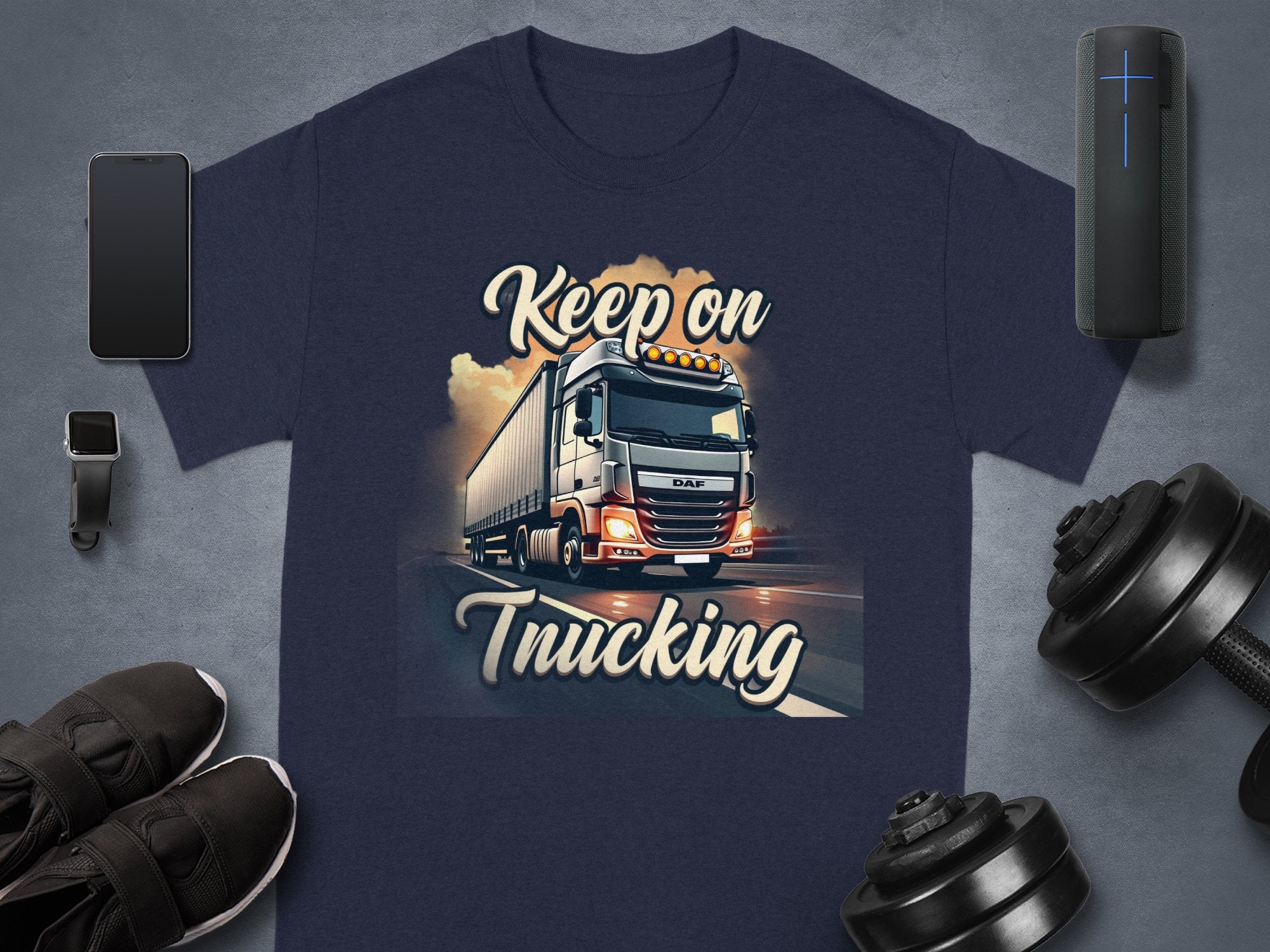 A Garment Graphics navy T-shirt featuring a semi-truck graphic and Keep on Trucking text is perfect for truck fans. Its surrounded by a smartphone, smartwatch, speaker, shoes, and a dumbbell on a gray surface.