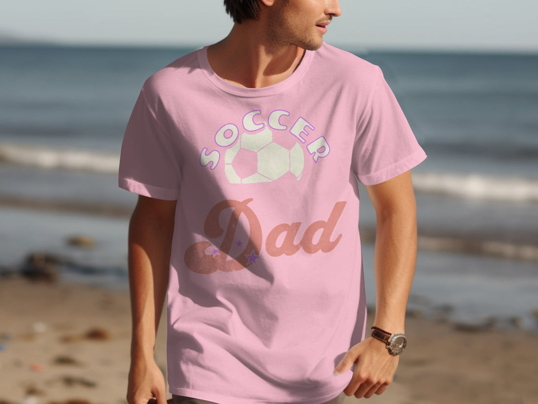 A Garment Graphics black T-shirt features Soccer Dad in stylized letters and a soccer ball graphic. Its displayed on a sports surface with a basketball, black shorts, water bottle, and white towel. Ideal for sporty dads balancing basketball on the court and soccer on the field.