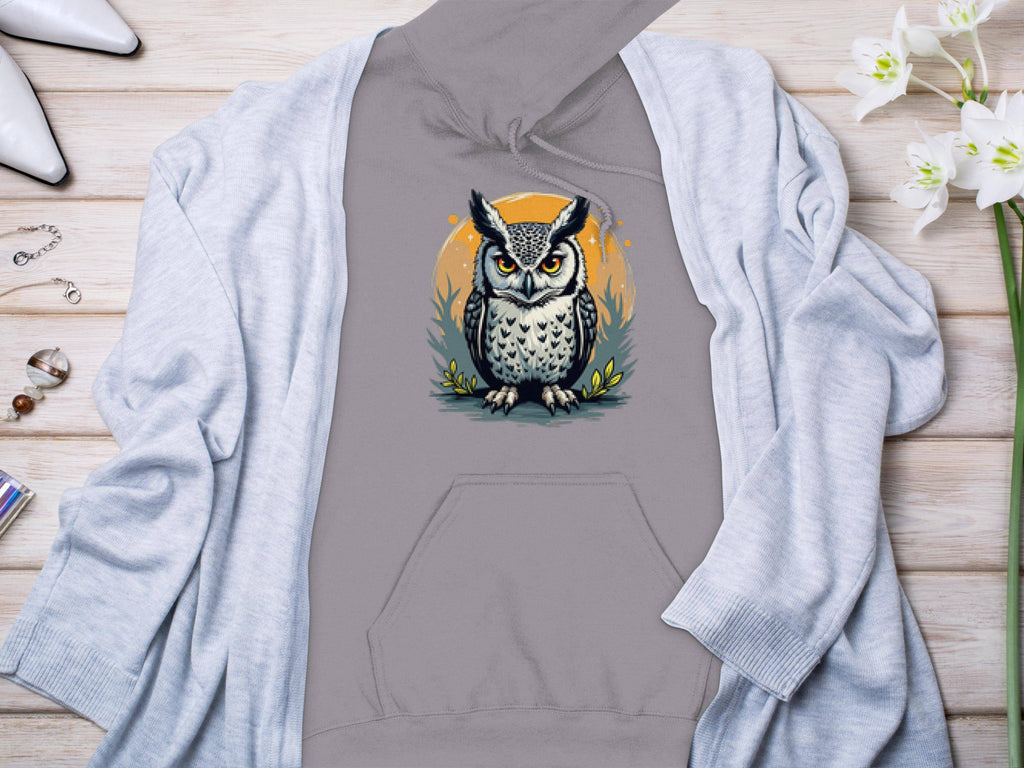 A Garment Graphics classic fit hoodie featuring detailed owl artwork lies beneath a light gray cardigan on a wooden surface. Nearby are white high heels, rings, a bracelet, and a bouquet of white flowers, merging elegance with comfort.
