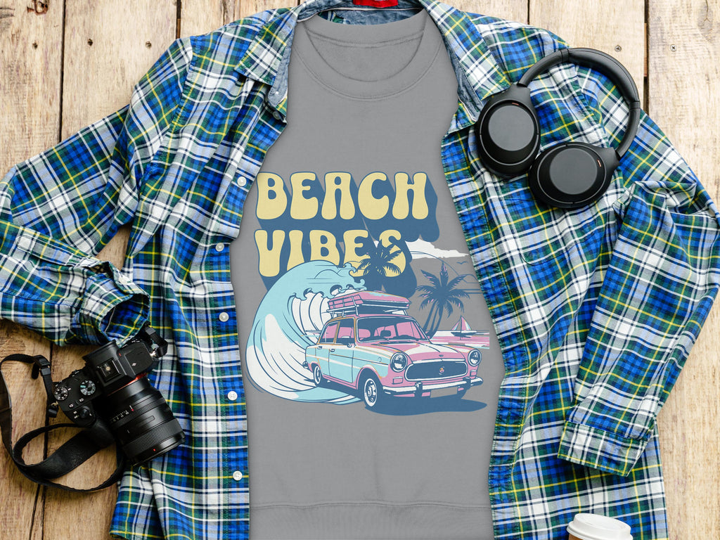 A unisex gray t-shirt by Garment Graphics features Beach Vibes text and a retro road trip image with a car, surfboards, palm trees, and waves. Its styled on wood with a plaid shirt, headphones, camera, and takeaway coffee cup.