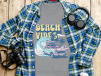 A unisex gray t-shirt by Garment Graphics features Beach Vibes text and a retro road trip image with a car, surfboards, palm trees, and waves. Its styled on wood with a plaid shirt, headphones, camera, and takeaway coffee cup.