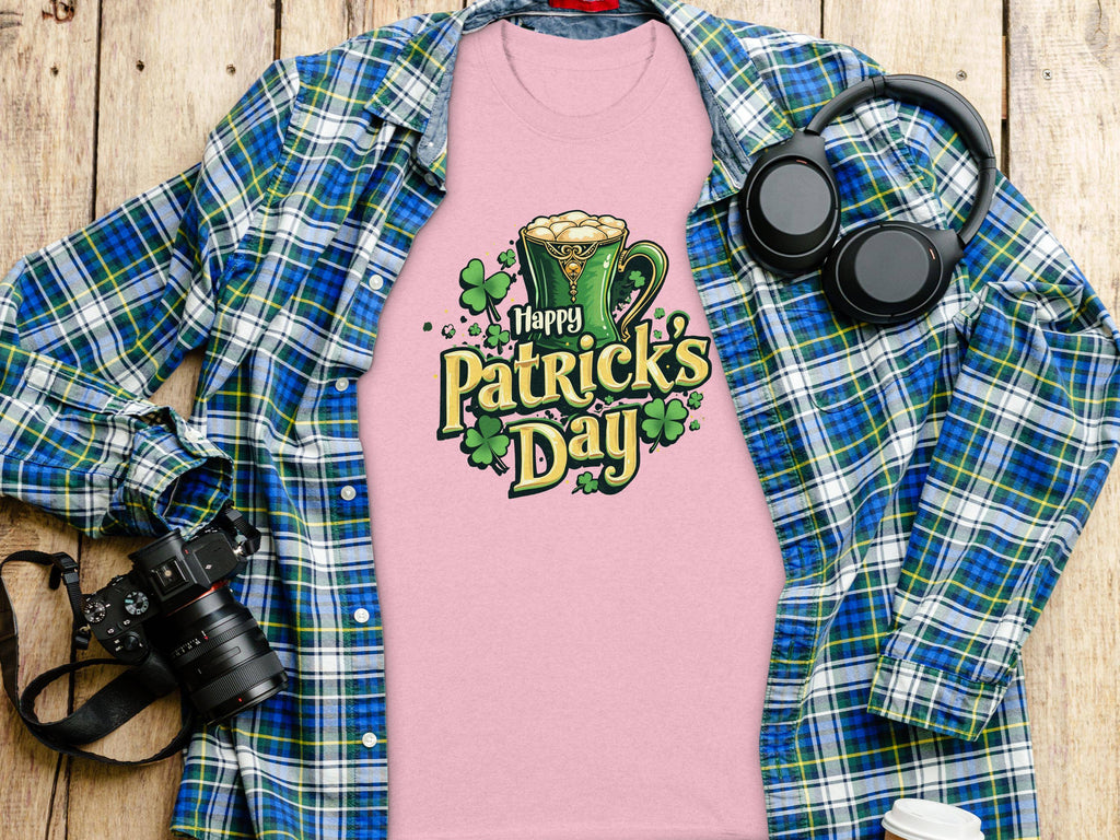 A statement pink Happy Patricks Day T-shirt by Garment Graphics, adorned with a leprechaun hat and clovers, rests on a wooden surface, surrounded by festive items: a blue and green plaid shirt, headphones, a camera, and a white beverage container.
