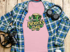 A statement pink Happy Patricks Day T-shirt by Garment Graphics, adorned with a leprechaun hat and clovers, rests on a wooden surface, surrounded by festive items: a blue and green plaid shirt, headphones, a camera, and a white beverage container.