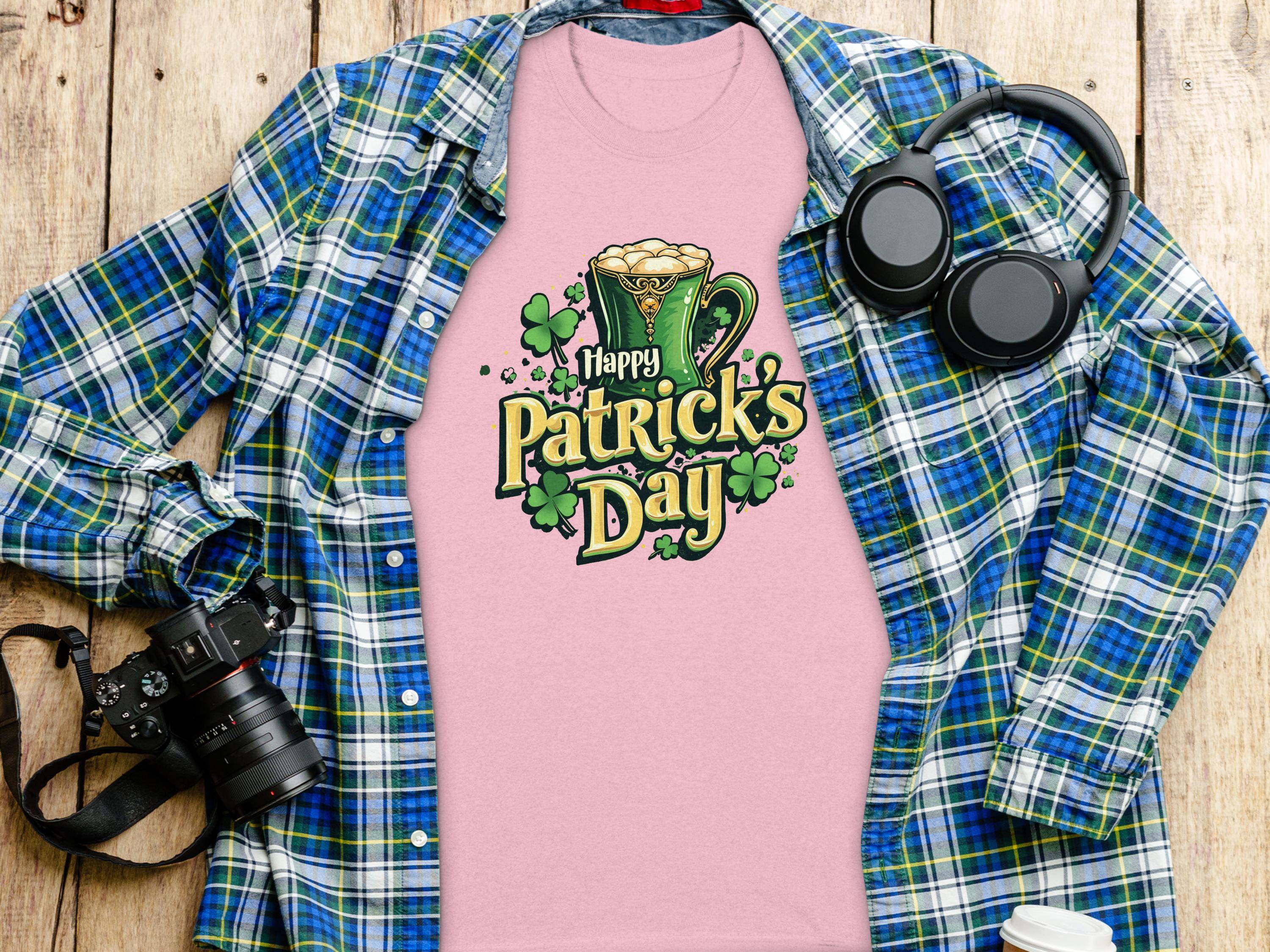 A statement pink Happy Patricks Day T-shirt by Garment Graphics, adorned with a leprechaun hat and clovers, rests on a wooden surface, surrounded by festive items: a blue and green plaid shirt, headphones, a camera, and a white beverage container.