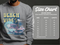 A person is wearing a Garment Graphics gray graphic sweatshirt with a vintage car, surfboards, palm trees, and Beach Vibes, ideal for a retro roadtrip. The Gildan 18000 size chart on the right shows unisex sizing from S to 5XL with measurements in inches.